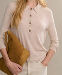 A woman with blonde wavy hair, face unseen, wears the Grayson Pullover—a light beige casual long-sleeve shirt—paired with cream pants and holds a large mustard-yellow suede clutch under her arm.