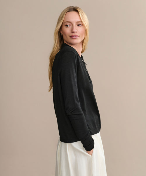 A woman with long blonde hair wears the Grayson Pullover, a black casual polo sweater, paired with a white skirt. She stands with her hands in her pockets, looking to the side against a plain beige background.
