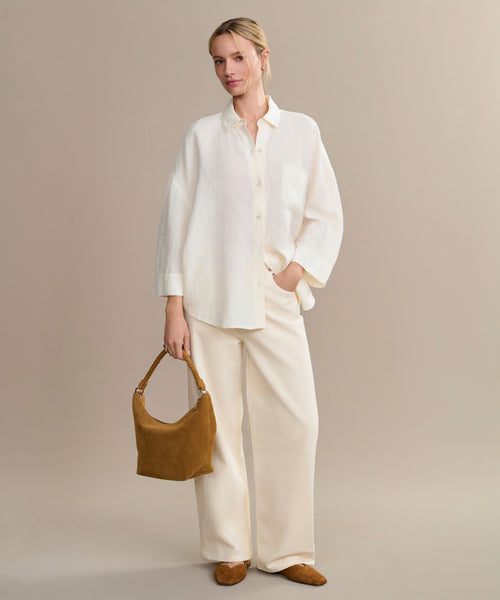A woman poses against a neutral background in the boxy-fit white Linen Logan Shirt and wide-leg cream pants, holding a tan suede handbag. She wears matching flats with her hair pulled back, exuding relaxed and effortless style.
