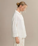 A blonde-haired woman in a low bun models the boxy-fit Linen Logan Shirt with matching pants, standing sideways against a simple beige background for an effortlessly stylish look.