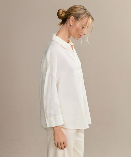 A blonde-haired woman in a low bun models the boxy-fit Linen Logan Shirt with matching pants, standing sideways against a simple beige background for an effortlessly stylish look.