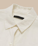 A close-up of the Linen Logan Shirt—a cream boxy-fit button-up with a pointed collar, white buttons, and a black label at the neck—laid flat on a neutral background.