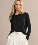 A woman with long blonde hair wears the Grayson Pullover in black, tucked into high-waisted white pleated pants. She stands against a plain beige background, gazing at the camera with a neutral expression.