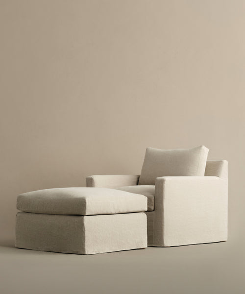 The Harbor Chair in Drift Linen, from Jenni Kayne Furniture, features clean lines and plush cushions. Paired with a matching ottoman, its modern cream design offers cozy comfort and minimalist style against a simple beige backdrop.