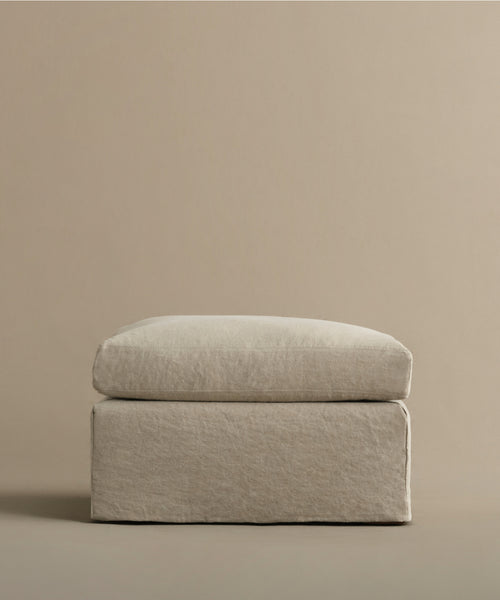 The Harbor Ottoman features a minimalist, square linen design with a cushioned top and clean lines, set against a matching beige background—a refined reflection of Jenni Kayne Furniture’s timeless style.