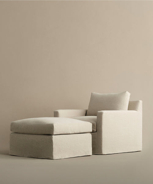 A modern, minimalist armchair with the Harbor Ottoman from Jenni Kayne Furniture, both upholstered in light beige linen and set against a plain, neutral background.