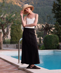 A woman in a wide-brimmed hat, white tank top, and the Kinney Skirt stands by a pool outdoors, surrounded by greenery and mountain views—capturing a perfect summer vibe.