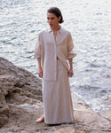 A woman stands barefoot on rocks by the water, wearing a loose beige button-up shirt and the Kinney Skirt, with waves and stones in the background.