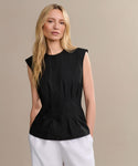 A woman with long blonde hair wears the Lara Top, a sleeveless black Italian washed poplin button-front shirt, paired with white pants. She stands against a plain beige background, looking relaxed and neutral.