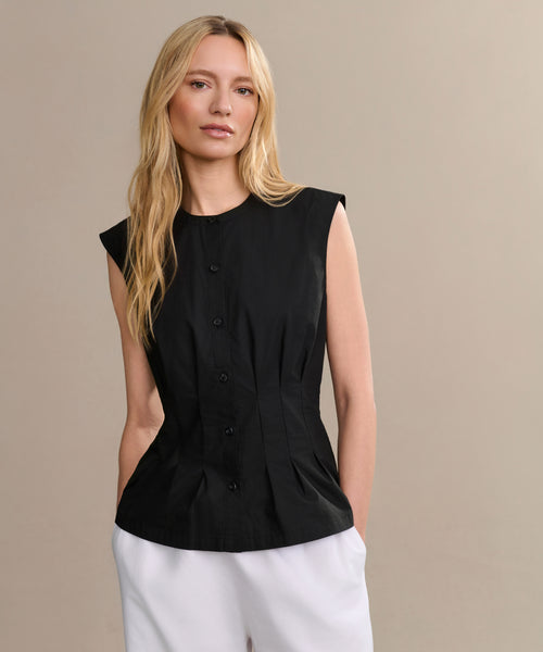 A woman with long blonde hair wears the Lara Top, a sleeveless black Italian washed poplin button-front shirt, paired with white pants. She stands against a plain beige background, looking relaxed and neutral.
