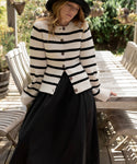 A woman in a striped sweater, Lauren Skirt, and hat leans against a wooden table on an outdoor patio surrounded by plants.