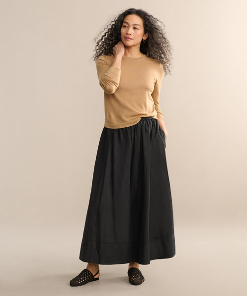 A woman with long curly hair stands against a plain background wearing a tan long-sleeve top, black slip-on shoes, and the Lauren Skirt in black cotton, with one hand tucked in her skirt pocket.