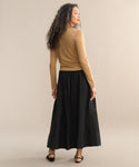 A woman with curly hair stands facing away against a neutral background, wearing a tan long-sleeve top, black sandals, and the Lauren Skirt—a chic black skirt.