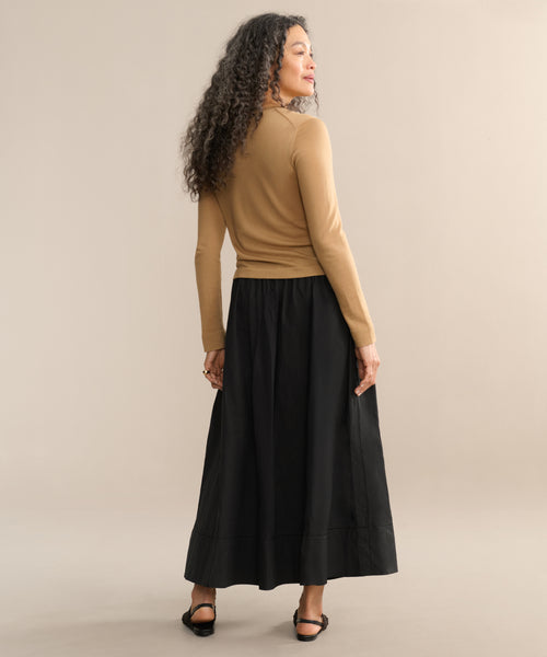 A woman with curly hair stands facing away against a neutral background, wearing a tan long-sleeve top, black sandals, and the Lauren Skirt—a chic black skirt.