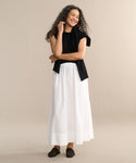 A woman with long curly hair smiles against a beige background. She wears a black sleeveless top, the wardrobe staple white Lauren Skirt—an ankle-length cotton style—and dark woven shoes.