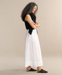 A woman with curly hair stands in profile against a plain background, wearing a black sleeveless top, the Lauren Skirt as her wardrobe staple, a black sweater draped over her shoulders, and black woven sandals.