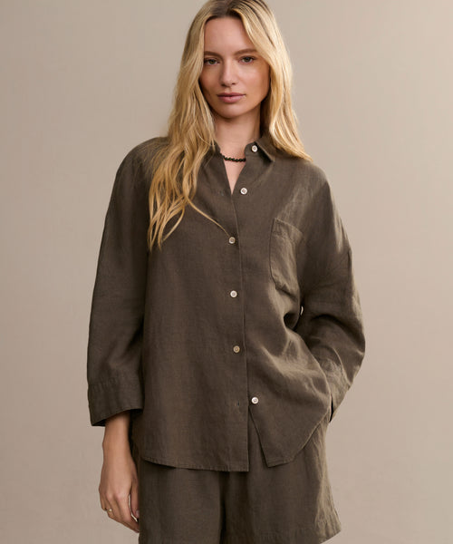 A woman with long blonde hair wears the dark olive Linen Logan Shirt and matching shorts in a boxy fit, standing against a plain beige background with one hand in her pocket for an effortlessly stylish look.