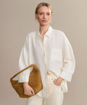 A woman stands against a beige background wearing the Linen Logan Shirt and light pants, holding a tan suede handbag with one hand in her pocket, effortlessly showcasing chic style.