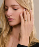 A woman with long blonde hair gently touches her face, showcasing the Liv Rose Cut Ring with a clear gemstone on her ring finger. Her calm expression and black top create a modern, elegant contrast.