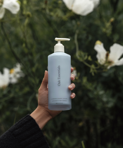 A hand in a black sleeve holds a light blue bottle from the Blue Tansy Bundle with a pump dispenser—formulated with blue tansy for dewy skin—set against blurred green foliage and white flowers.