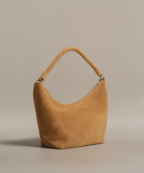 The Medium Crescent Bag in camel-colored suede features a curved design and thick twisted handle, crafted in Italy. Photographed on a plain beige background.