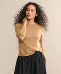 A woman with curly hair models the Mercer Crewneck, styled with a black skirt. She stands against a beige background, gazing to the side with one hand at her neck and the other in her pocket.