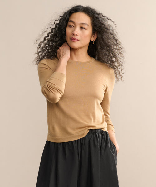 A woman with curly hair models the Mercer Crewneck, styled with a black skirt. She stands against a beige background, gazing to the side with one hand at her neck and the other in her pocket.