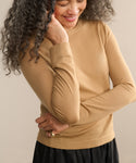A smiling woman with curly gray hair looks down, wearing a tan Mercer Crewneck sweater and a black skirt. Her right hand touches her hair while her left arm rests across her waist.