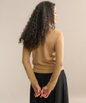 A woman with long, curly hair stands facing away, hands clasped behind her back. She wears the Mercer Crewneck sweater in a classic cut and a black skirt, set against a plain beige background.