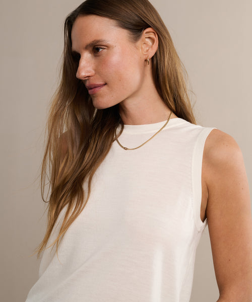 A woman with long brown hair wears the Merino Tank, featuring a sleeveless white design, paired with a gold necklace and small hoop earrings as she looks to the side against a neutral beige background.