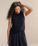 A woman with long curly hair smiles while looking down, wearing the Merino Tank in black with a matching skirt, one hand touching her hair, standing against a plain beige background.
