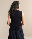 A person with long, curly hair wears a sleeveless black Merino Tank and a dark skirt, standing with their back to the camera against a plain beige background.