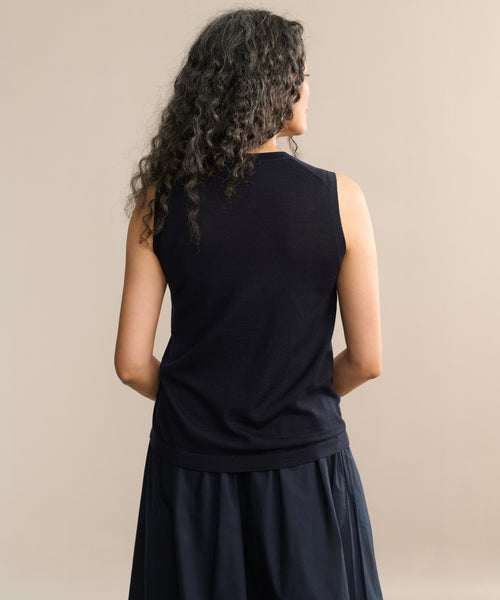 A person with long, curly hair wears a sleeveless black Merino Tank and a dark skirt, standing with their back to the camera against a plain beige background.