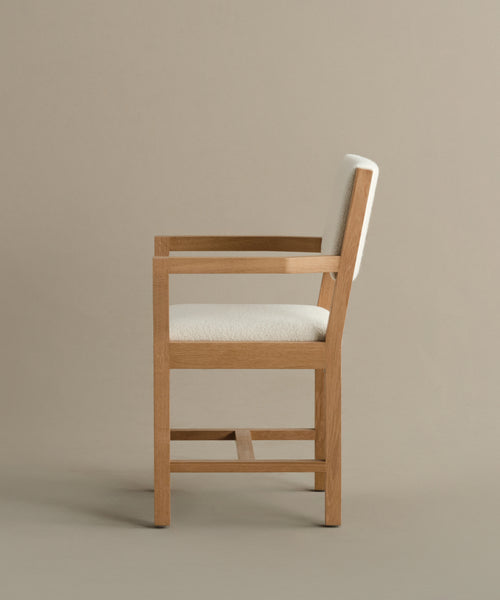 Side view of the Montecito Upholstered Dining Arm Chair with a light-colored cushioned seat and backrest, clean straight lines, and minimalist white oak design, set against a plain beige background.