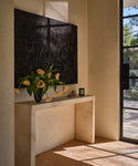 A minimalist entryway features the Napa Console Table in ivory, topped with a vase of yellow tulips and small decor items, complemented by a large black abstract artwork and sunlight streaming through a glass door.