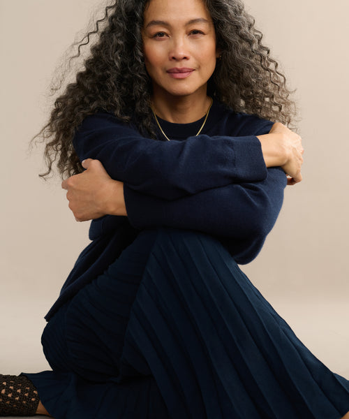A woman with long, curly gray hair smiles softly while sitting on the floor in a Pleated Skirt and sweater, her arms wrapped around her knees against a plain background.