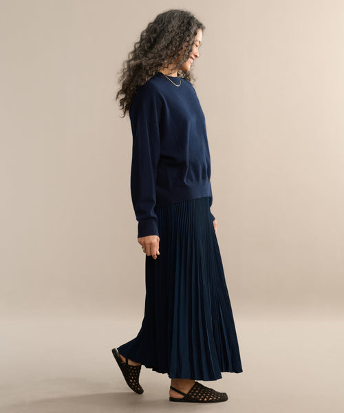 A smiling woman with curly hair models a pleated skirt and a navy sweater, walking barefoot on a beige background, facing left.
