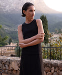 A woman with short dark hair wears a sleeveless black Cotton Rib Tank and skirt, standing outdoors with arms crossed in front of a stone wall and a scenic mountain backdrop.