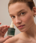 A person with fair skin holds up a green bottle of Blue Tansy Bundle oil with a dropper cap, promising dewy skin and rich hydrators, while looking directly at the camera against a softly lit, plain background.