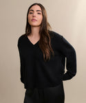 A woman with long brown hair wears the Simone V-Neck Black sweater, crafted from fine cotton merino, and a black skirt against a beige background—showcasing this perfect everyday layer.