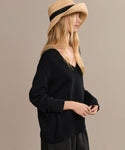 A woman in the Simone V-Neck, a loose black sweater made from fine cotton merino, stands against a beige background. She wears a wide-brimmed straw hat and her long blonde hair flows as she looks to the side with hands gently together.