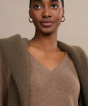 A person wears the Simone V-Neck sweater in brown cotton merino with a textured brown jacket over their shoulders and gold hoop earrings, set against a neutral background.