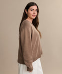 A woman with long brown hair wears the Simone V-Neck, a loose cotton-merino sweater, with white pants. She stands sideways, looking at the camera against a plain beige background—an effortless luxury layer.