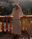 A woman in the Simone V-Neck ivory fine cotton merino sweater and long skirt stands by a stone railing, sunlight behind her, with trees and hills beyond—a perfect luxury layer for any season.