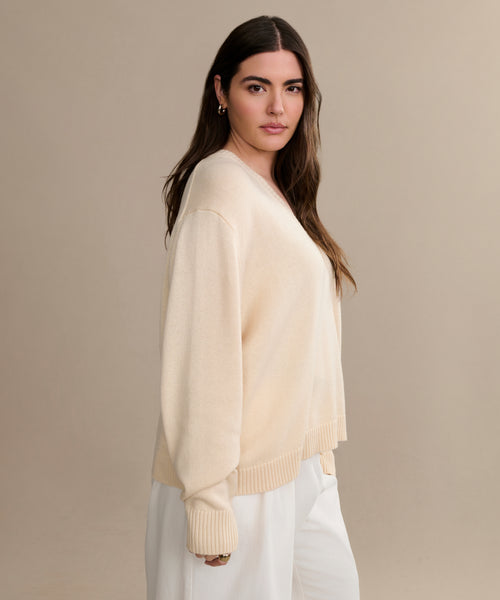 A woman with long brown hair wears the Simone V-Neck in Ivory fine cotton merino and white pants, standing against a beige background with a neutral expression—a luxurious, versatile layer for any occasion.