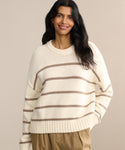 A woman with long dark hair smiles, wearing the Chloe Crewneck—a cream Peruvian cotton sweater with brown stripes—and tan pants, standing against a plain beige background.