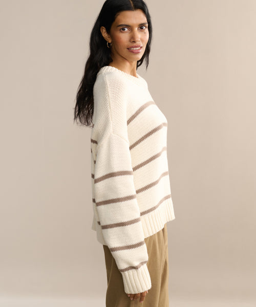 A woman with long dark hair wears the Chloe Crewneck, a cream sweater with brown stripes, paired with tan pants against a beige background. She smiles gently at the camera, highlighting the cozy cropped fit.