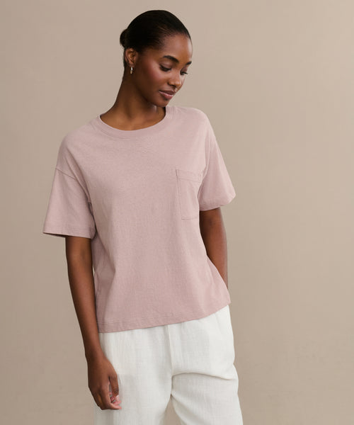 Wearing the Vintage Pocket Tee in light pink with a chest pocket and white pants, this look against a beige background and subtle smile makes it an ideal everyday wardrobe choice.