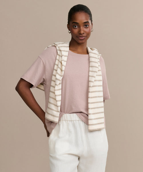 A woman wears the Vintage Pocket Tee in light pink, paired with white pants and a cream-beige striped sweater over her shoulders, standing against a beige background and smiling softly with one hand in her pocket.