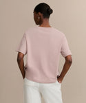A person stands with hands on hips, wearing the Vintage Pocket Tee in light pink and white pants. Their hair is styled in a neat bun with hoop earrings, creating an effortless casual look against a plain beige background.